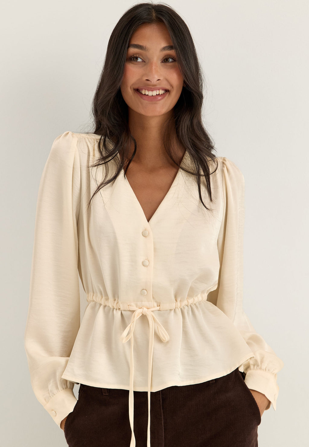 Kermanvärinen satiinipusero edestä, V-pääntie, sävyyn sopivat napit ja vyötärön kiristysnyöri.